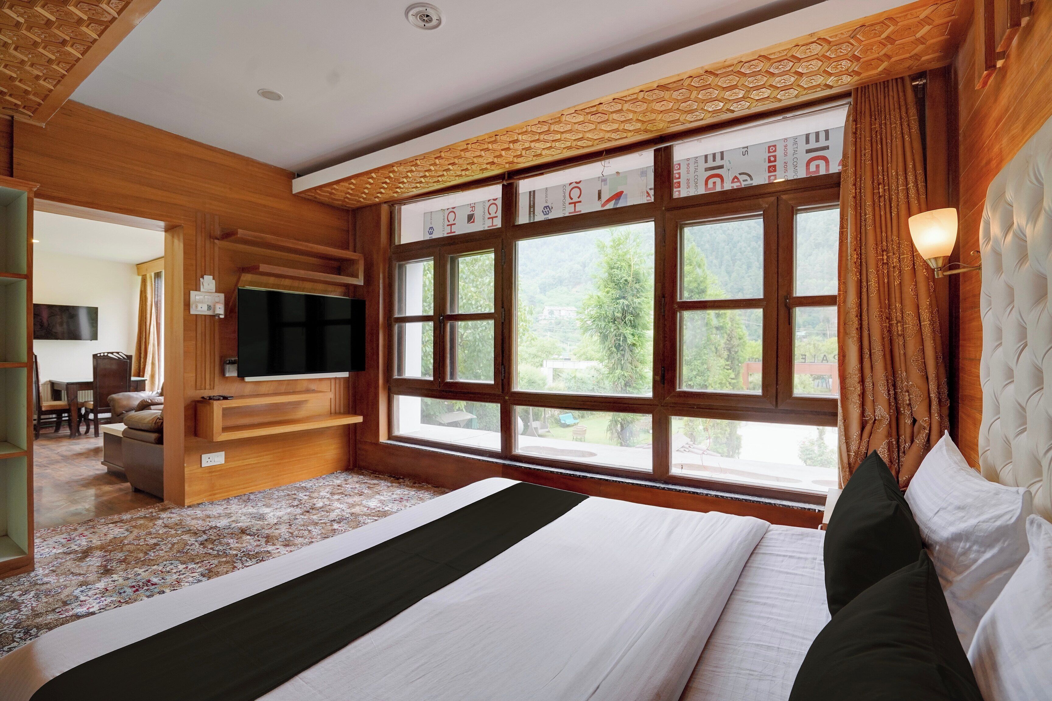 A bedroom with a large bed, a television, a window with curtains, and a wooden ceiling.
