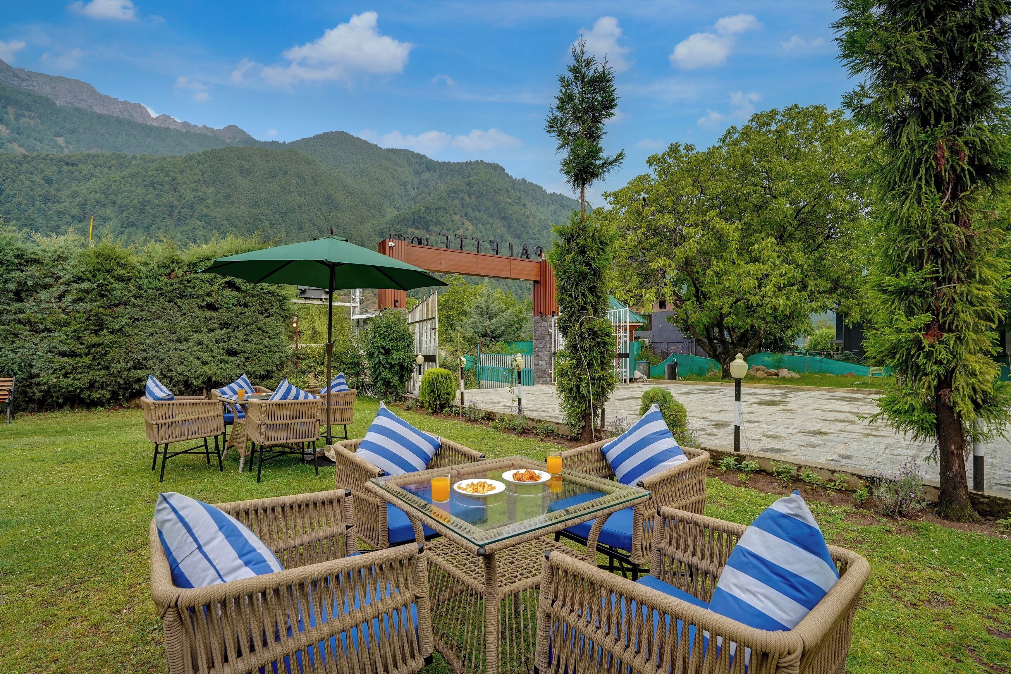 An outdoor seating area with wicker furniture and a glass table, surrounded by greenery and a mountain backdrop.