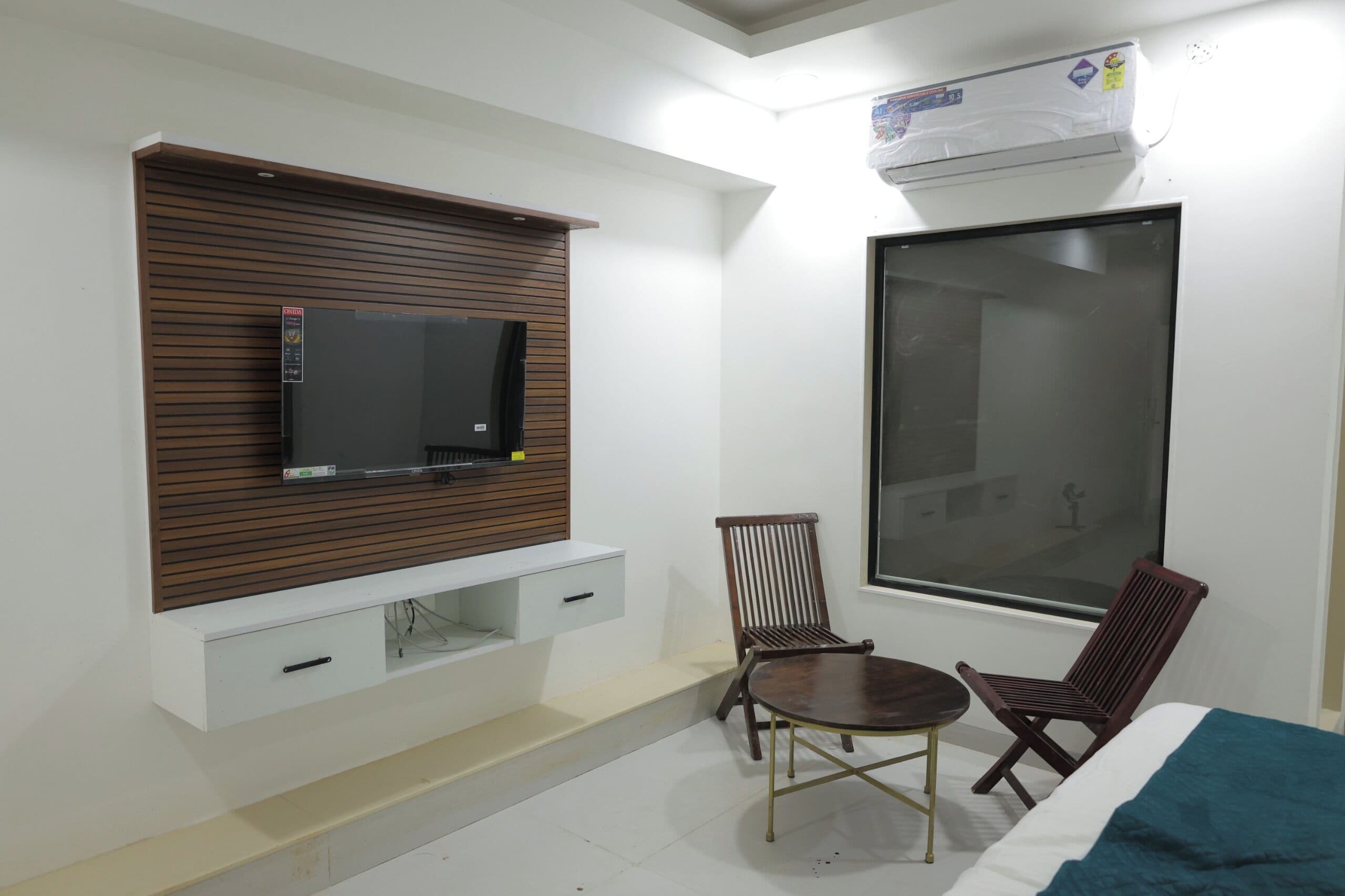 Una habitación con televisión montada en un panel de madera, una ventana, una cama con manta azul y dos sillas de madera.