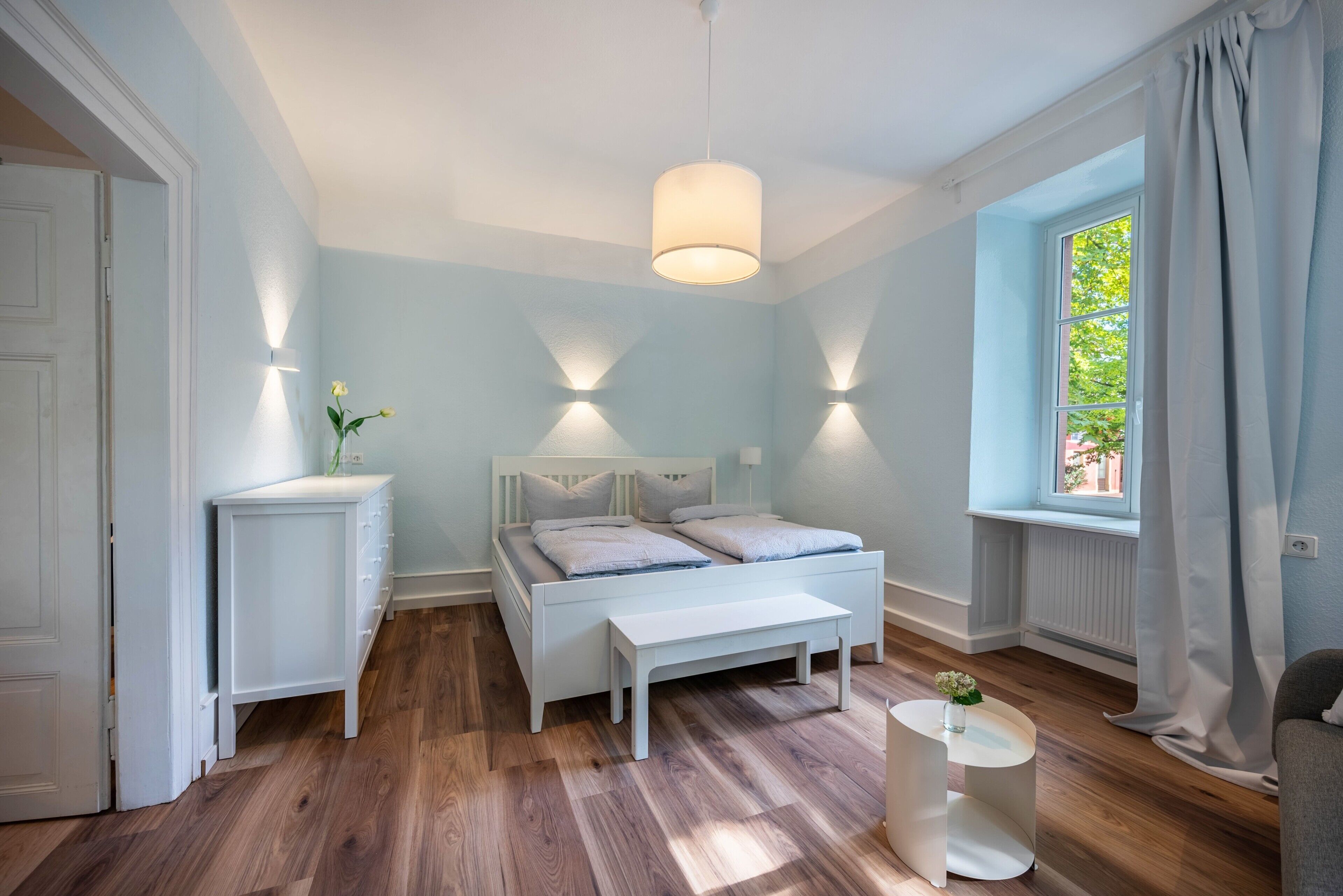 A bedroom with a bed, bedside tables, a bench, a window, and a hanging light fixture.