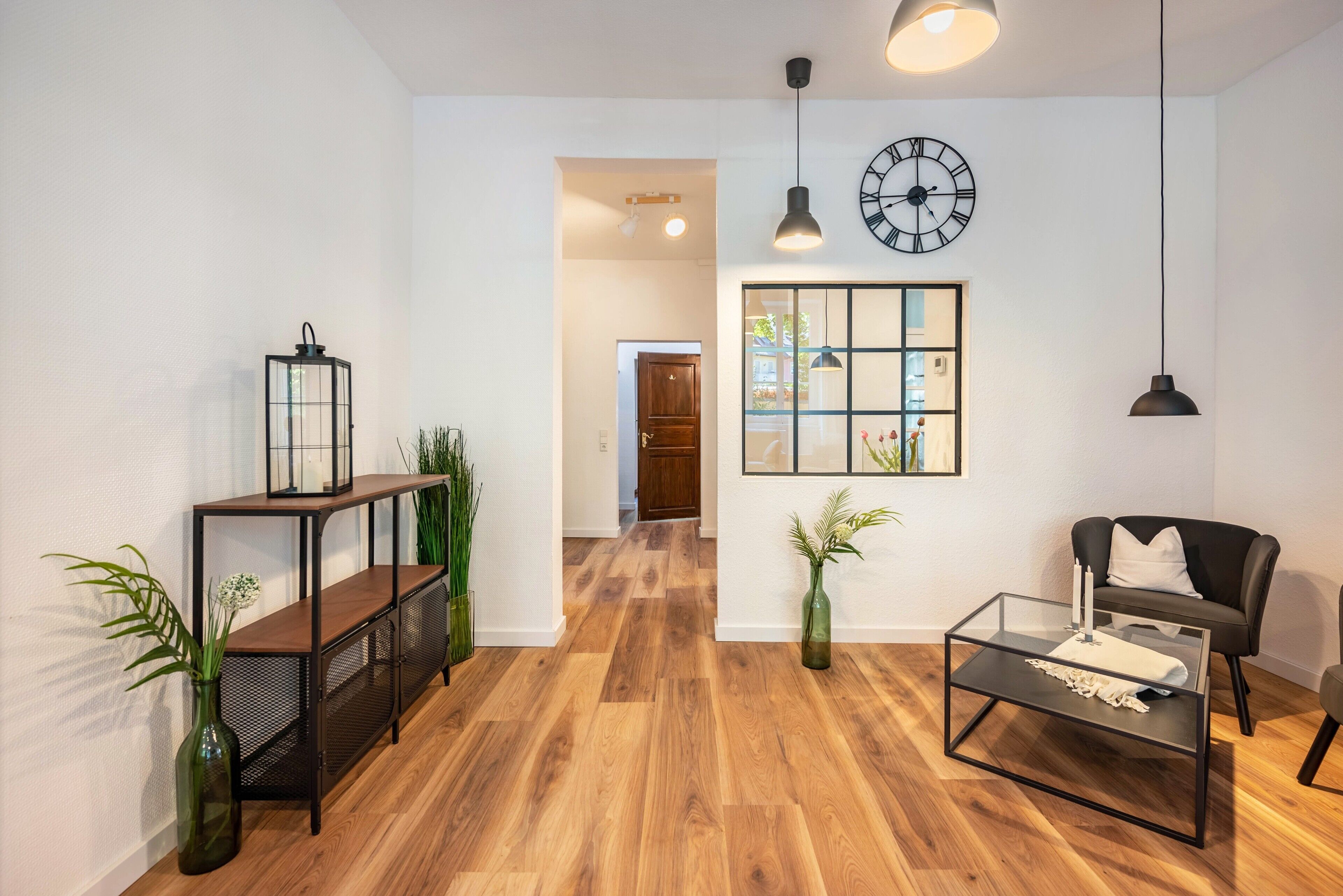Ein modernes Wohnzimmer mit Holzboden, einem Glastisch, einem schwarzen Sofa, einer Wanduhr und einem großen Fenster mit Blick.