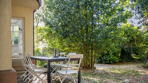 En udendørs patio med et bord og stole, omgivet af grøn vegetation og træer.