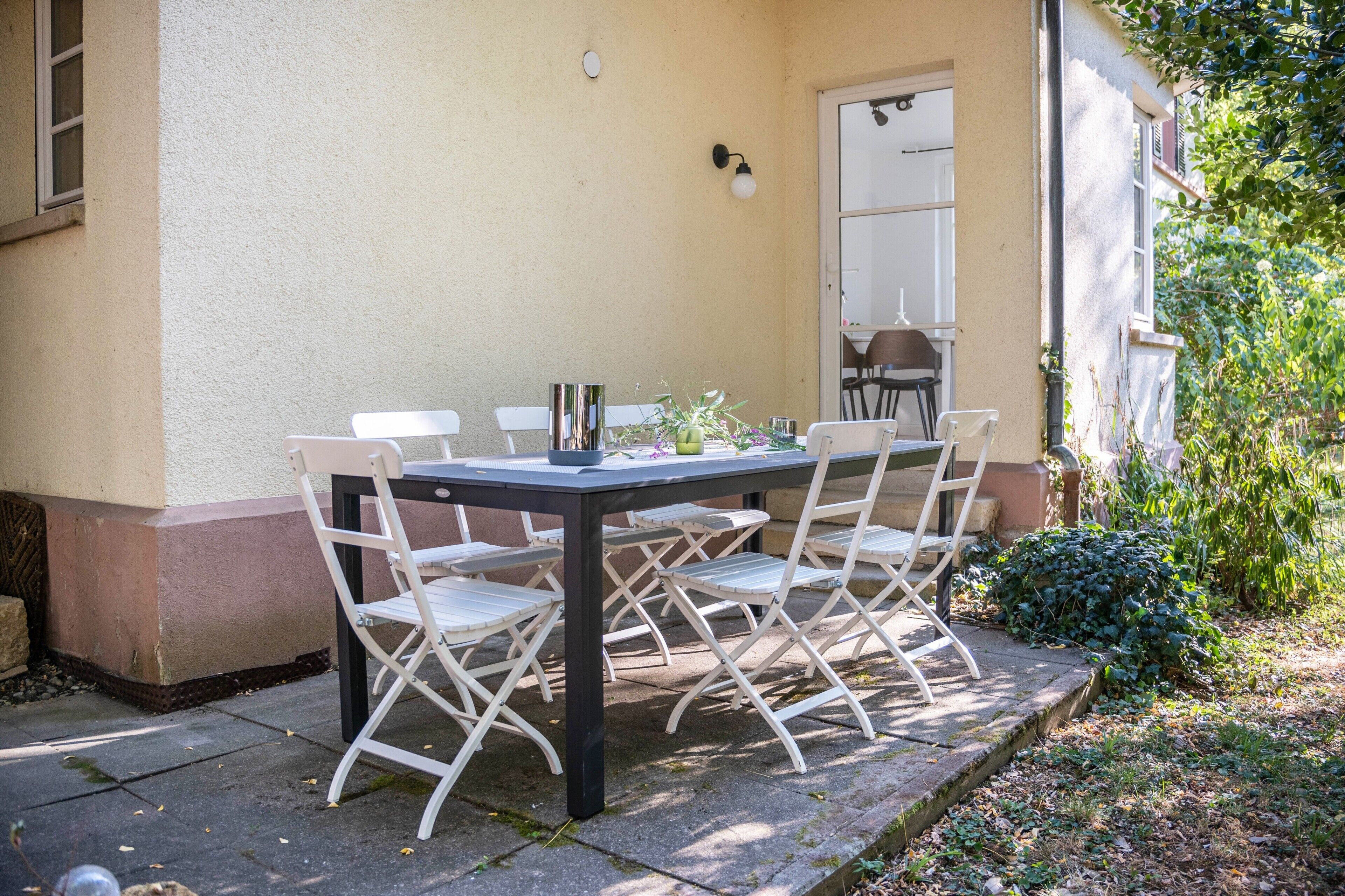 Une terrasse extérieure avec une table et des chaises, un luminaire fixé au mur et une porte donnant sur un espace intérieur.
