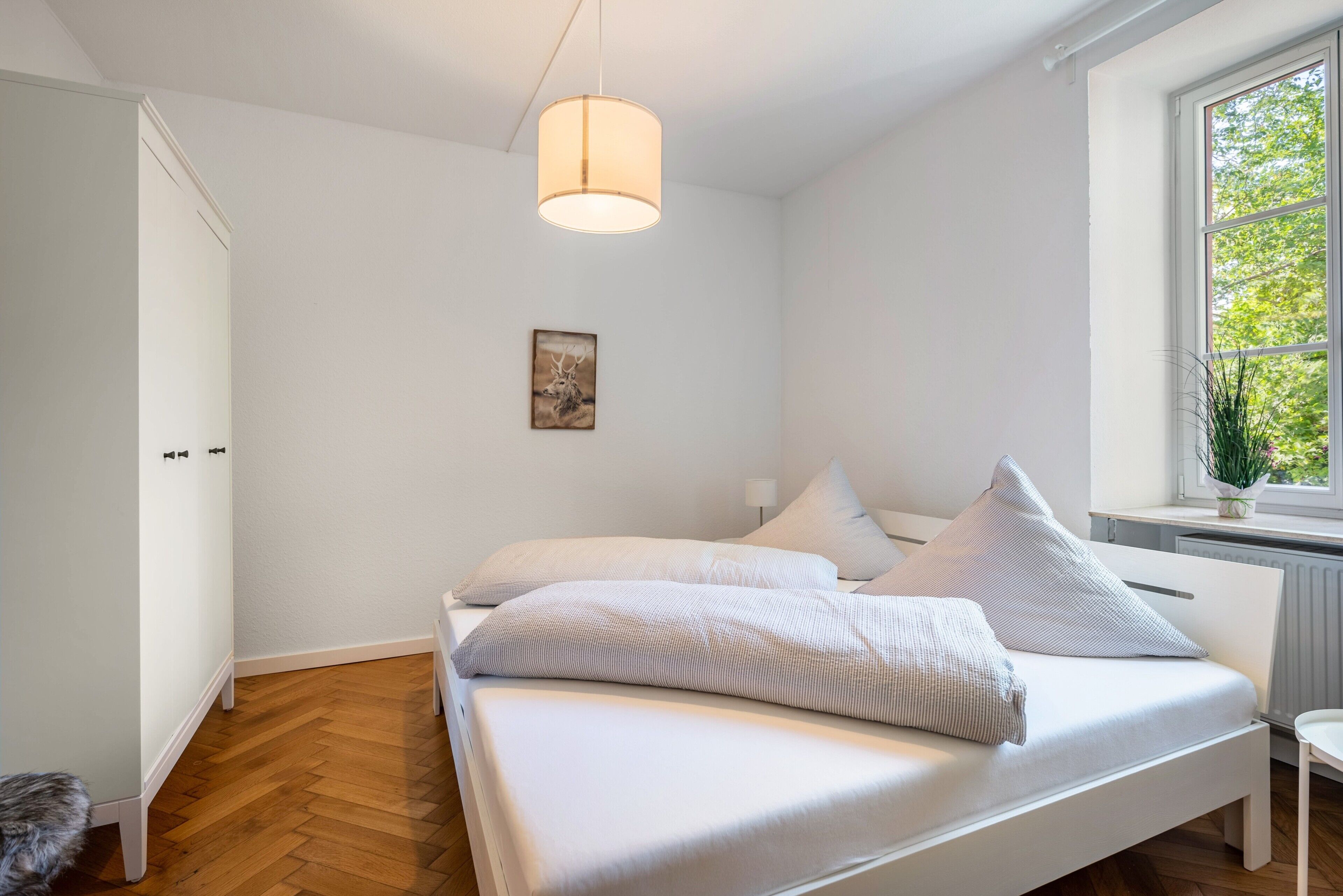 Ein Schlafzimmer mit einem Bett, einer Pendelleuchte, einem Fenster mit Blick ins Grüne und einem Bild an der Wand.