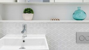 Et badeværelse med en hvid vask, et spejl, en potteplante, en glasflaske og en toiletpapirholder.