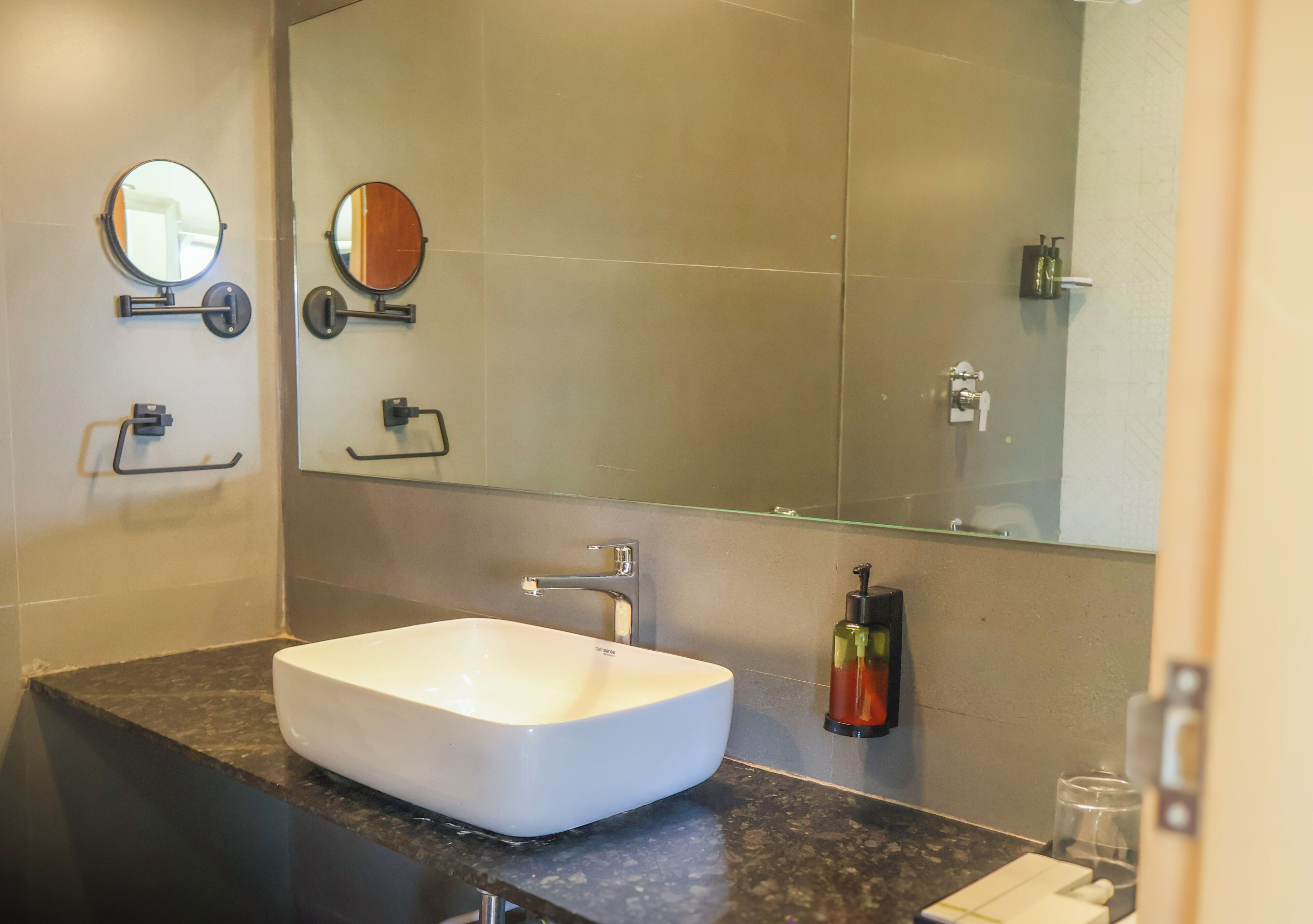 Un baño con un lavamanos blanco, dos espejos en la pared, un espejo grande y un recipiente de vidrio sobre la encimera.