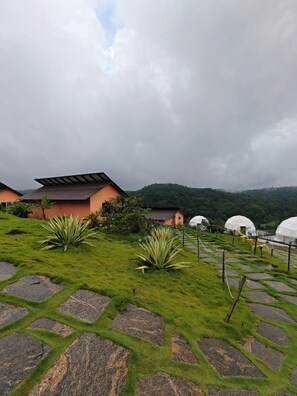 A terraced garden with stone pathways, a building with a sloped roof, and a hill in the background. - The Divine Valley By Devandhari (Sakleshpur)