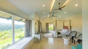 A bedroom with a four-poster bed, a sitting area with a chair and a small table, and a view of a lush landscape through large windows. - The Divine Valley By Devandhari (Sakleshpur)