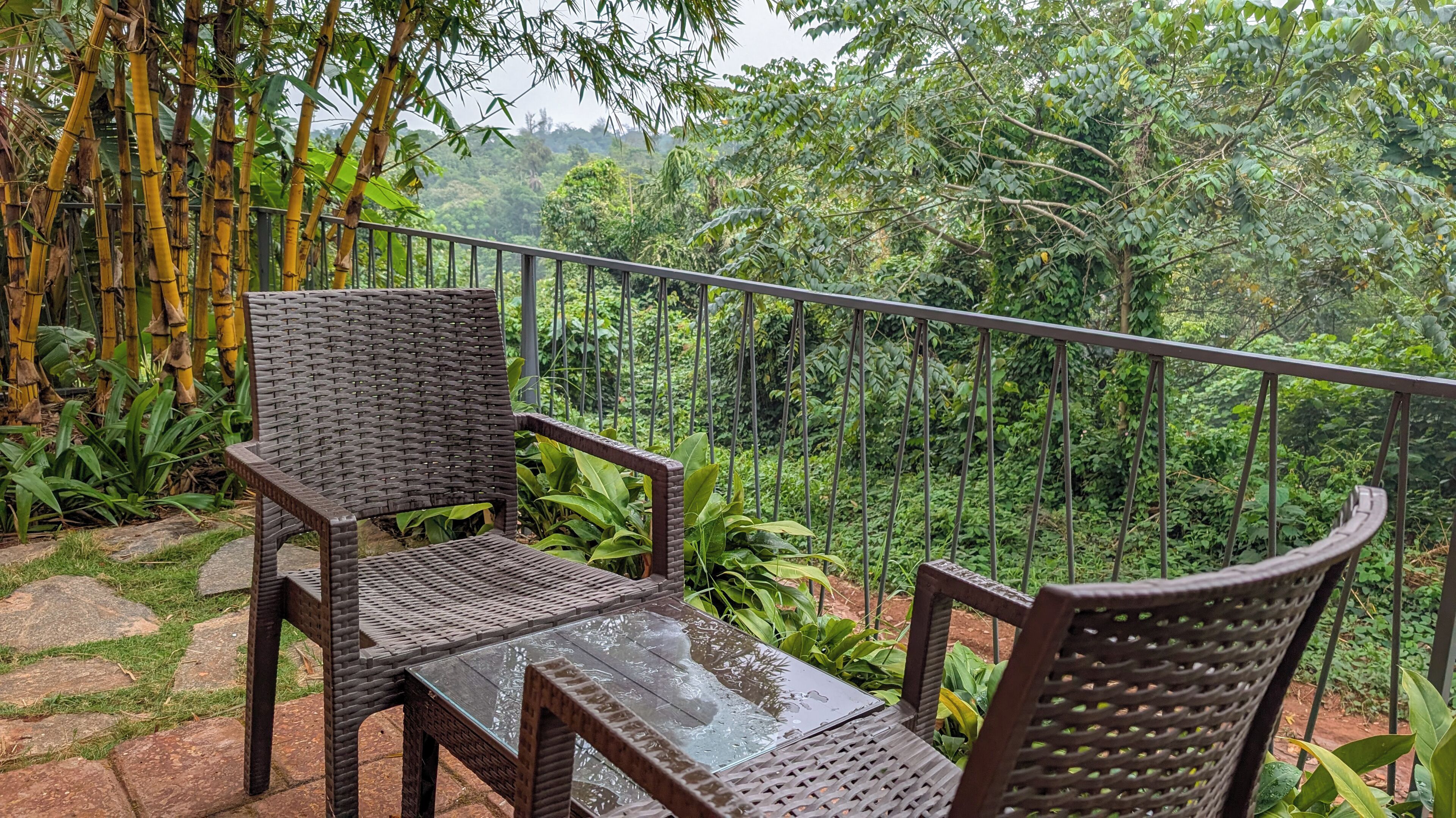 A balcony with wicker furniture and a glass table, surrounded by lush greenery and bamboo.