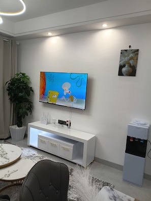 A modern living room with a flat-screen TV mounted on the wall, a white entertainment unit, a potted plant, a water dispenser, and a marble-patterned table. - Modern cozy Retreat (Nairobi)