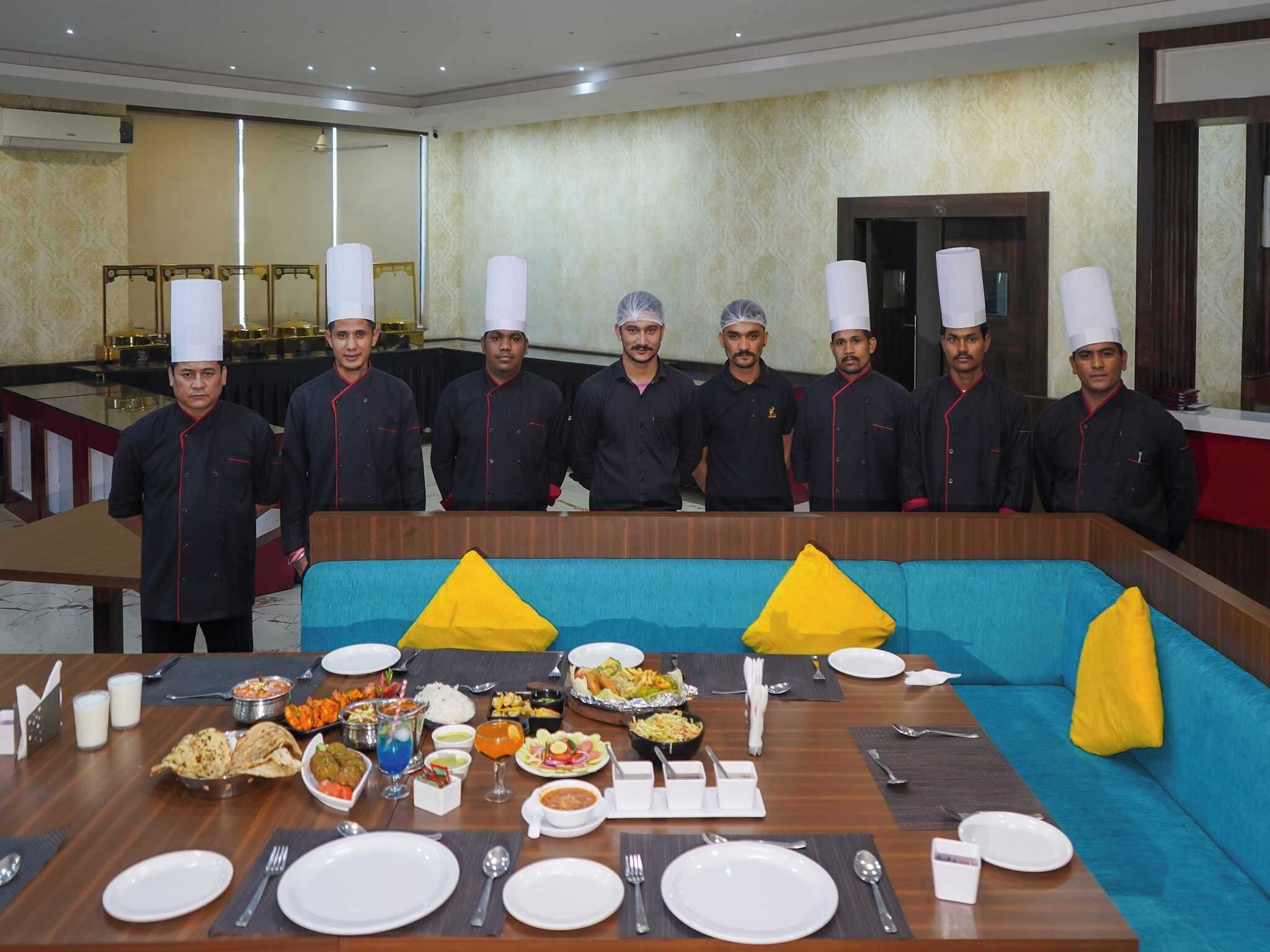 Un grupo de personal uniformado de servicio parado detrás de una mesa de comedor dispuesta con diversos platos y un sofá azul con cojines amarillos.