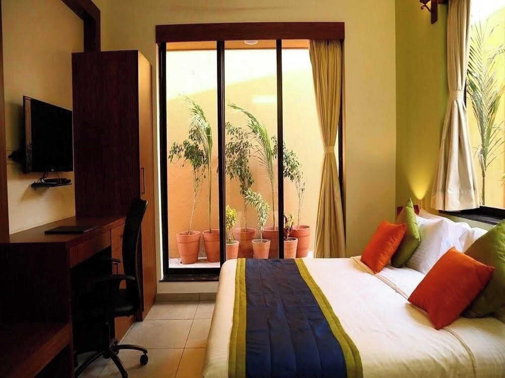 Habitación de hotel con cama, escritorio, silla, televisor y vista a un balcón con plantas en macetas.