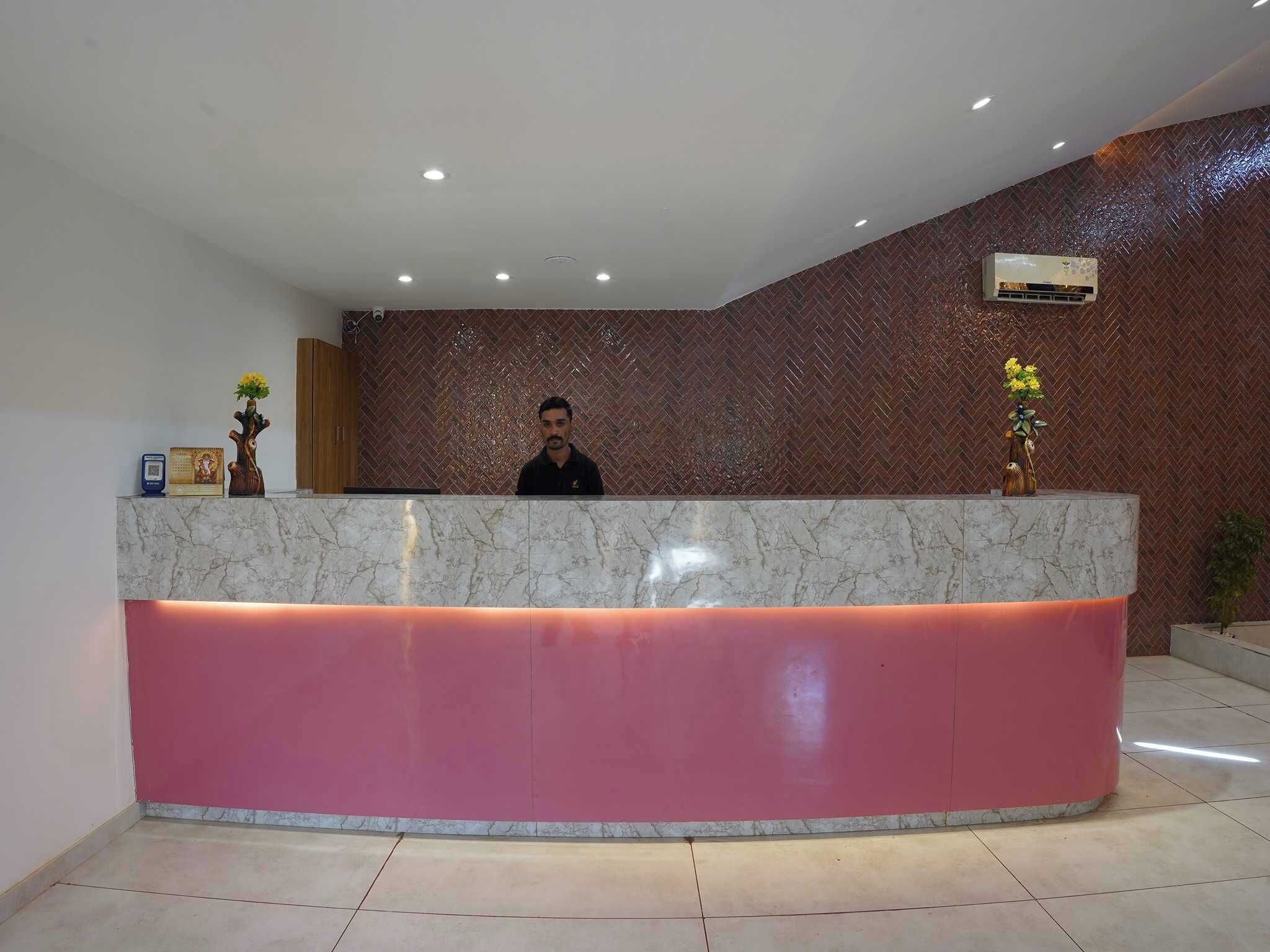 A reception desk with a marble top and pink base, a person standing behind it, and a herringbone-patterned wall with an air conditioning unit.