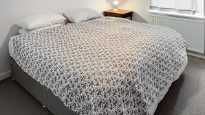 Uma cama com colcha estampada, uma mesinha de cabeceira com abajur e uma janela com cortinas.