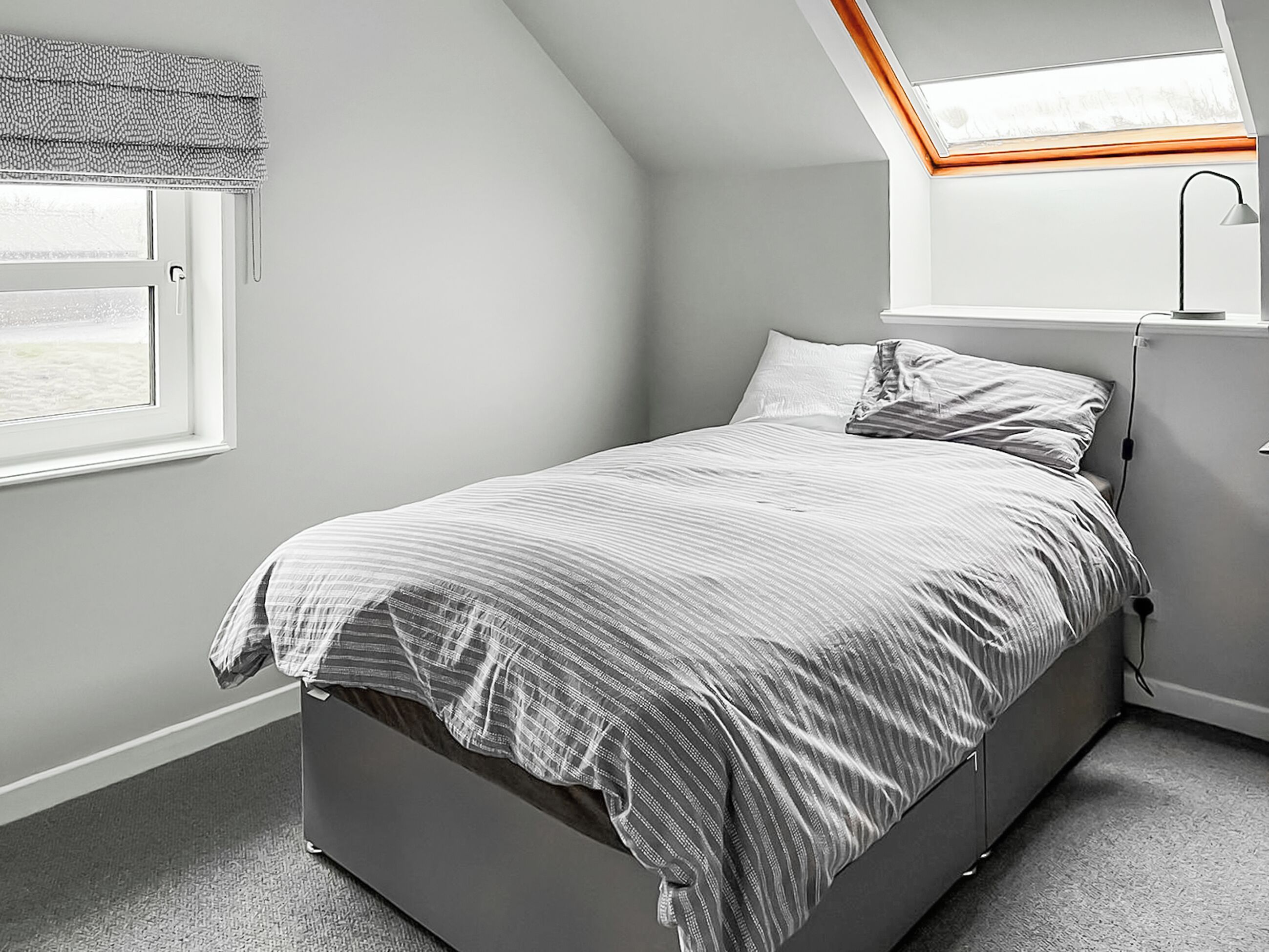 A bedroom with a bed, a skylight, a window, and a desk with a lamp.