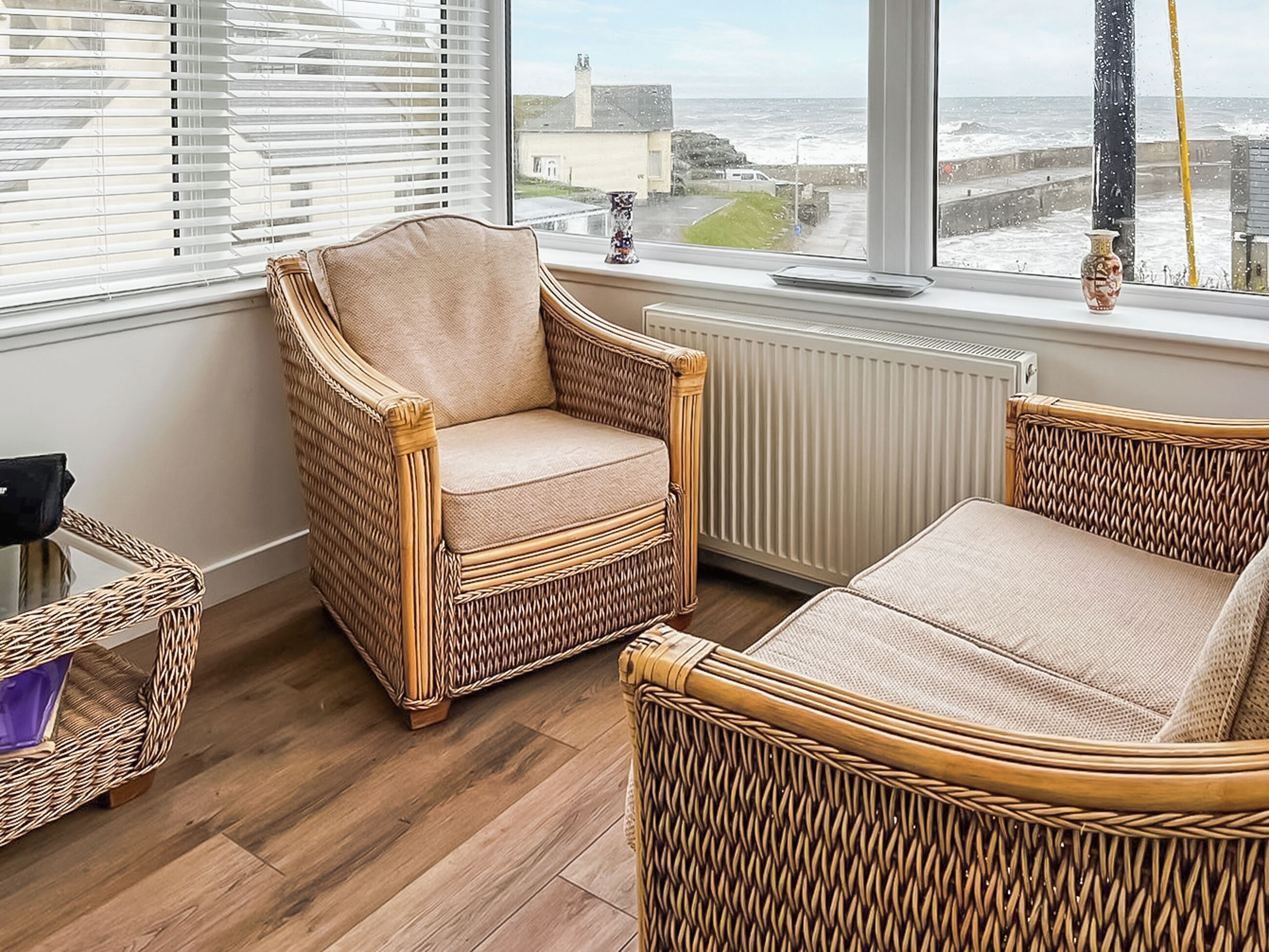 A room with a wicker chair, a glass table, and a view of the sea through large windows.