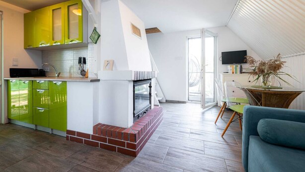 Una cocina moderna con armarios verdes, una chimenea de ladrillo y un comedor con una silla transparente y un sofá azul.