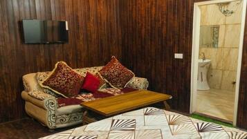 Una habitación con un revestimiento de madera, un sofá con cojines estampados, una mesa de centro y un baño visible a través de una puerta abierta.