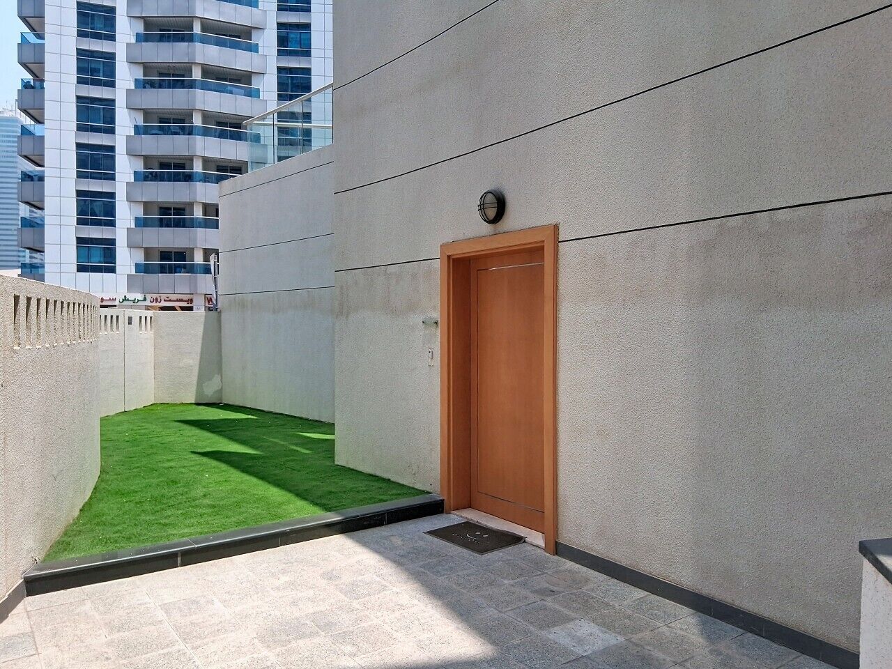 A narrow outdoor corridor with a wooden door, a small patch of artificial grass, and a modern building in the background.