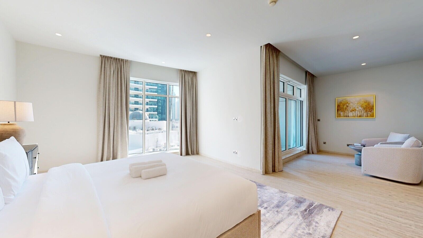 Ein Hotelzimmer mit einem großen Bett, einer Sitzecke mit einer Couch und Blick auf eine Stadtlandschaft durch große Fenster.