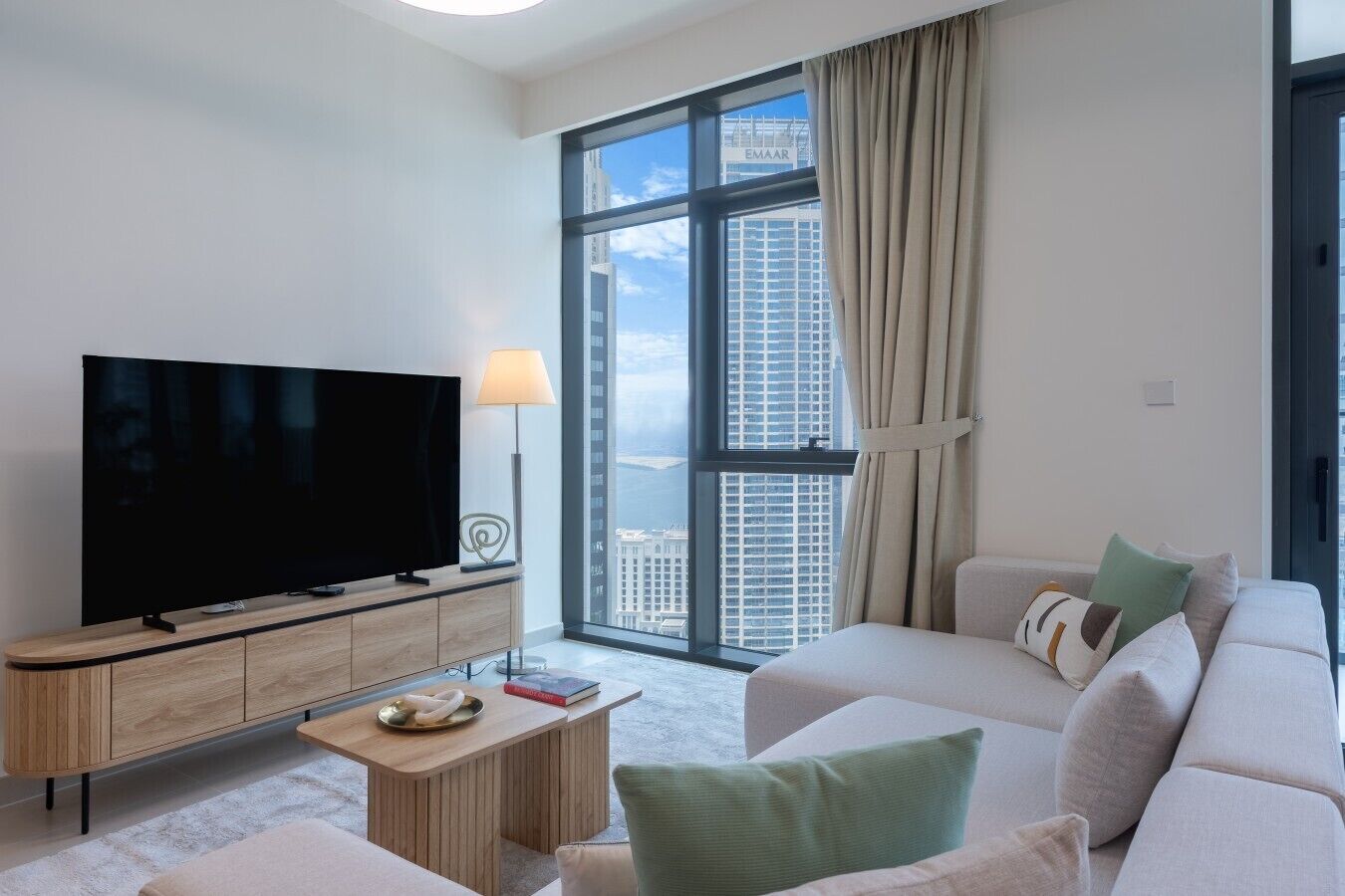 A modern living room with a flat-screen TV, a wooden coffee table, a sofa, and a large window with a view of high-rise buildings.