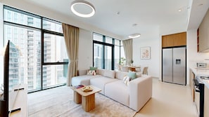 A modern living room with a sectional sofa, a coffee table, and a kitchen area with a refrigerator and stove. - Contemporary 2 Bed in Creek l Free WiFi & Pool (Creek Harbour)