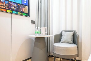 A modern hotel room with a flat-screen TV mounted on the wall, a small round table with a coffee maker and bottled water, a grey armchair, and a window with curtains. - Jinjiang Qingyang Qinglan Hotel (Quanzhou)
