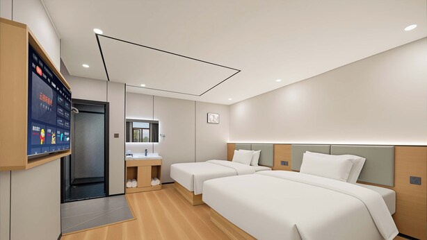 Una habitación de hotel moderna con dos camas, una pantalla digital mostrando información y un baño con espejo y lavamanos.