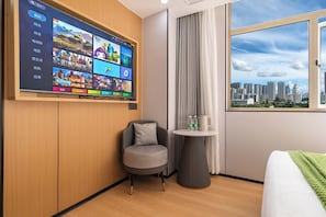 A hotel room with a large TV screen displaying various channels, a comfortable chair, a small table with a kettle and water bottles, and a view of the cityscape through a window. - Jinjiang Qingyang Qinglan Hotel (Quanzhou)