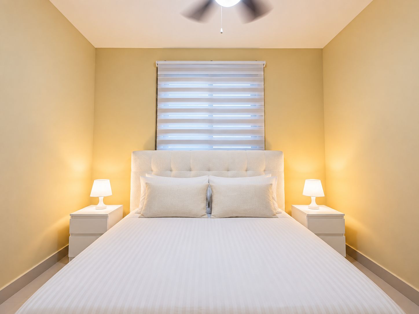 Une chambre à coucher avec un lit, deux tables de chevet, une fenêtre avec des stores et un ventilateur de plafond.