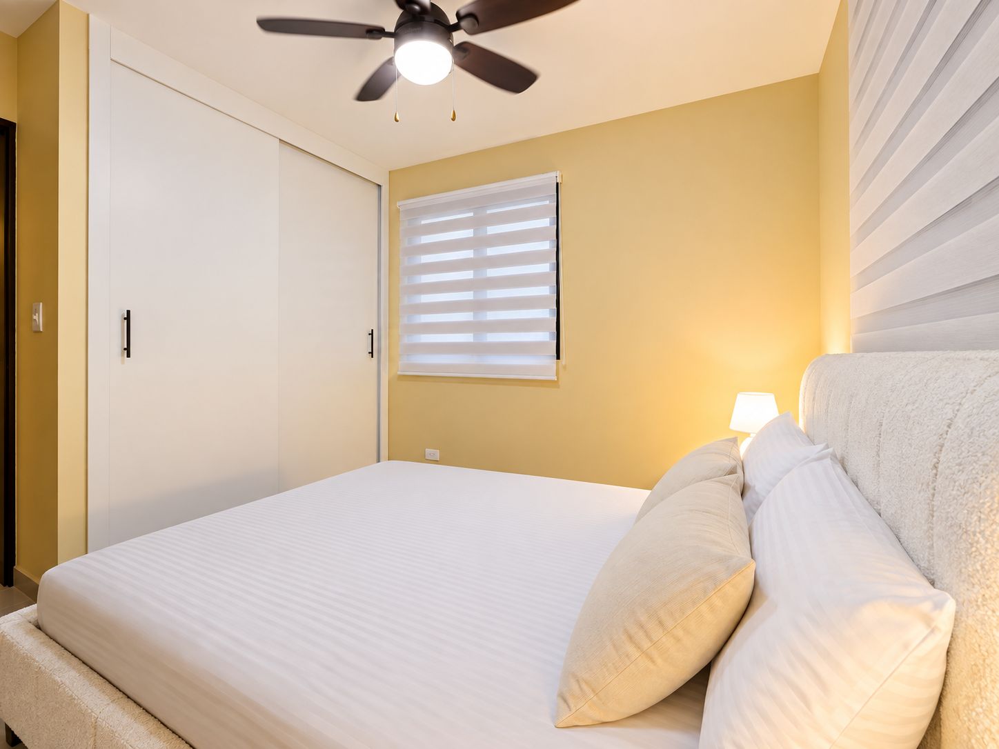 A bedroom with a bed, a ceiling fan, a window with blinds, and a bedside lamp.