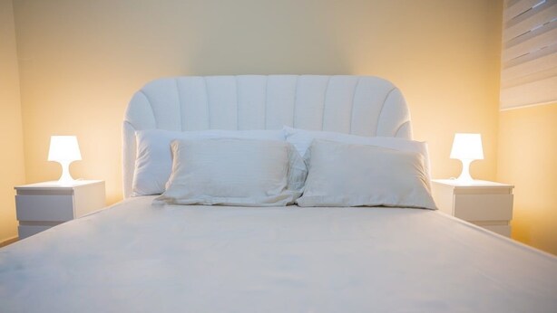Una cama con ropa de cama y almohadas blancas, flanqueada por dos mesitas de noche con lámparas, en una habitación con pared lisa y techo inclinado.