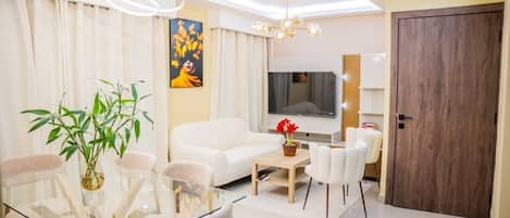 A modern living room with a sofa, coffee table, and chairs, a dining table with chairs, a large TV, and a wooden door.