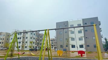 Un terrain de jeux avec des balançoires et un sol recouvert de petits graviers, avec un immeuble à plusieurs étages en arrière-plan.