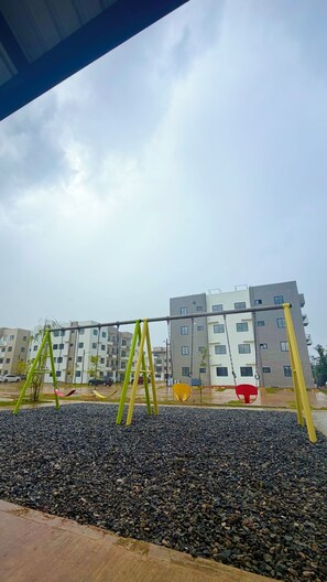 A playground with swings and a pebbled ground, with a multi-story building in the background. - Modern 3BR Apartment Near SDQ Airport with Free Parking, Fast Wi-Fi (Santo Domingo Este)