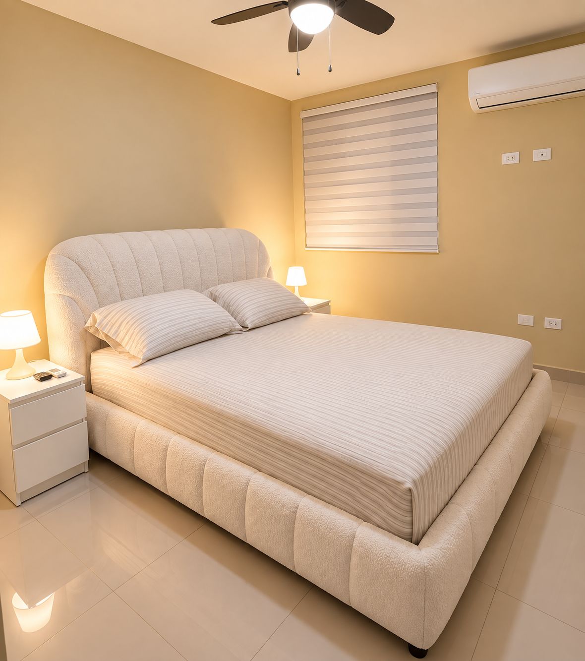 Une chambre à coucher comprenant un lit, des tables de chevet, un ventilateur de plafond, une fenêtre avec des stores et un appareil de climatisation.