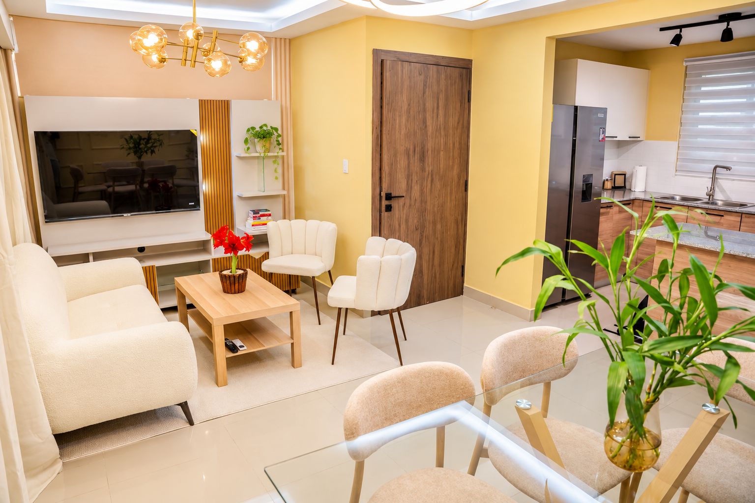 Un salon moderne avec un canapé, des fauteuils, une table basse et un téléviseur. On peut voir une porte en bois et un coin cuisine.