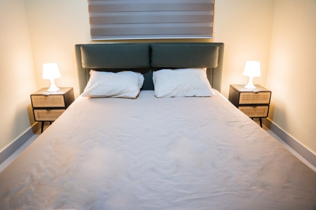 Ein ordentlich bezogenes Bett mit zwei Kissen, an beiden Seiten Flanken von Nachttischen mit Lampen, in einem Zimmer mit schlichtem, klaren Design.