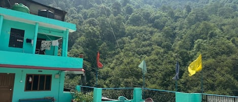 A two-story building with a flat roof, a balcony with chairs, and a view of a forested hillside.