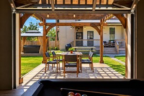 An outdoor patio area with a wooden pergola, a pool table, and a dining table with chairs.
