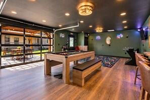 A pool table in a room with a view of a house through large windows, a neon sign, and a ceiling fan.