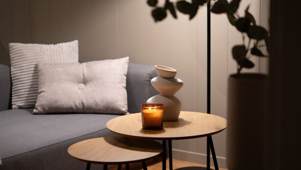 Un salon moderne avec un canapé gris, une petite table ronde en bois sur laquelle est posée une bougie, et un vase décoratif.