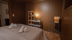 Une chambre d’hôtel avec un lit, deux serviettes enroulées, un bureau avec une lampe, une chaise et une étagère avec des livres et une plante.