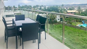 Varanda com guarda-corpo de vidro, mesa de jantar com cadeiras e vista para uma paisagem verde com piscina.