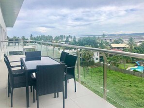 Ein Balkon mit Glastrennwand, ein Esstisch mit Stühlen und Blick auf eine grüne Landschaft mit Schwimmbad.
