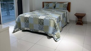 Um quarto com uma cama, uma pequena mesa de cabeceira de madeira, ar condicionado no teto e uma porta de vidro deslizante que dá para uma área externa.