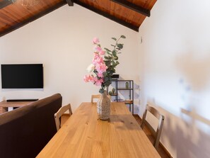 Un coin repas avec une table en bois, des chaises, un vase de fleurs, un téléviseur à écran plat et une étagère ornée d’un cactus.