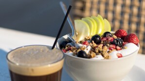A bowl of yogurt topped with fruits and granola, a glass of dark liquid, and a wicker chair in the background.