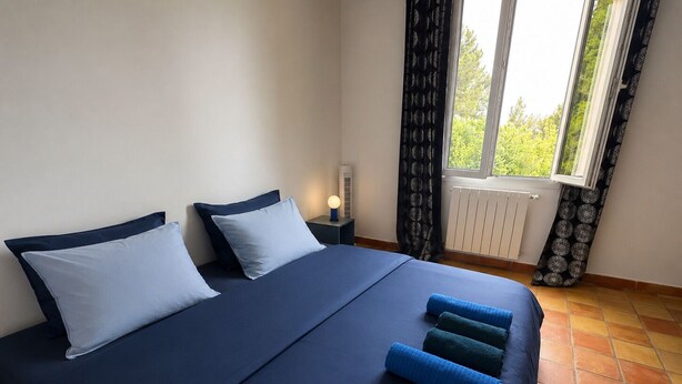 Un dormitorio con cama, almohadas, una mesita de noche, lámpara, ventana con cortinas y radiador.