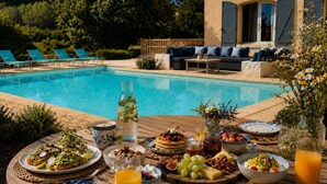 Desayuno junto a la piscina con diversas opciones de comida, un vaso de jugo de naranja y vistas a la piscina y la zona de estar al aire libre.